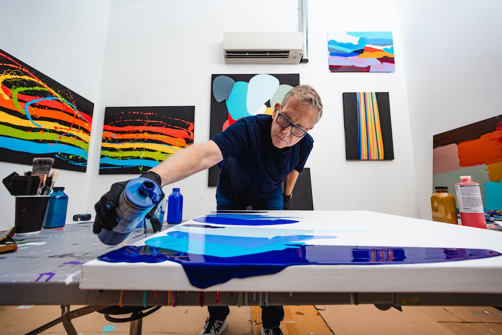 Photo of artist in studio standing before a new canvas, pouring blue paint down on it, by Austin-based lifestyle photographer Nick Barnes.