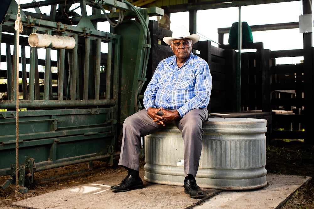 Portrait of African American rancher with sunglasses in cowboy hat and blue plaid button-down shirt, sitting on aluminum trough, by Tampa, Florida-based agricultural photographer Jeremiah Wilson.