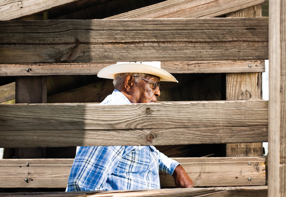 Image of African American rancher with sunglasses in cowboy hat and blue plaid button-down shirt, leaning on weathered wooden ranch frame, by Tampa, Florida-based agricultural photographer Jeremiah Wilson.
