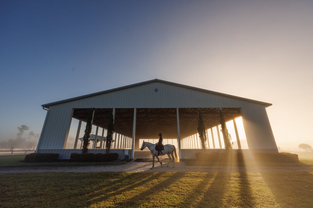 Profile of silhouetted rider on horseback, before open farm building,  in early morning sunlight with thick haze, by Fort Myers, FL-based lifestyle photographer Brian Tietz.