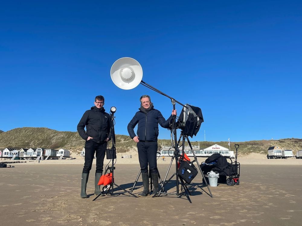 Behind the scenes portrait of photographer and his assistant with photo equipment on Dutch beach on clear day.