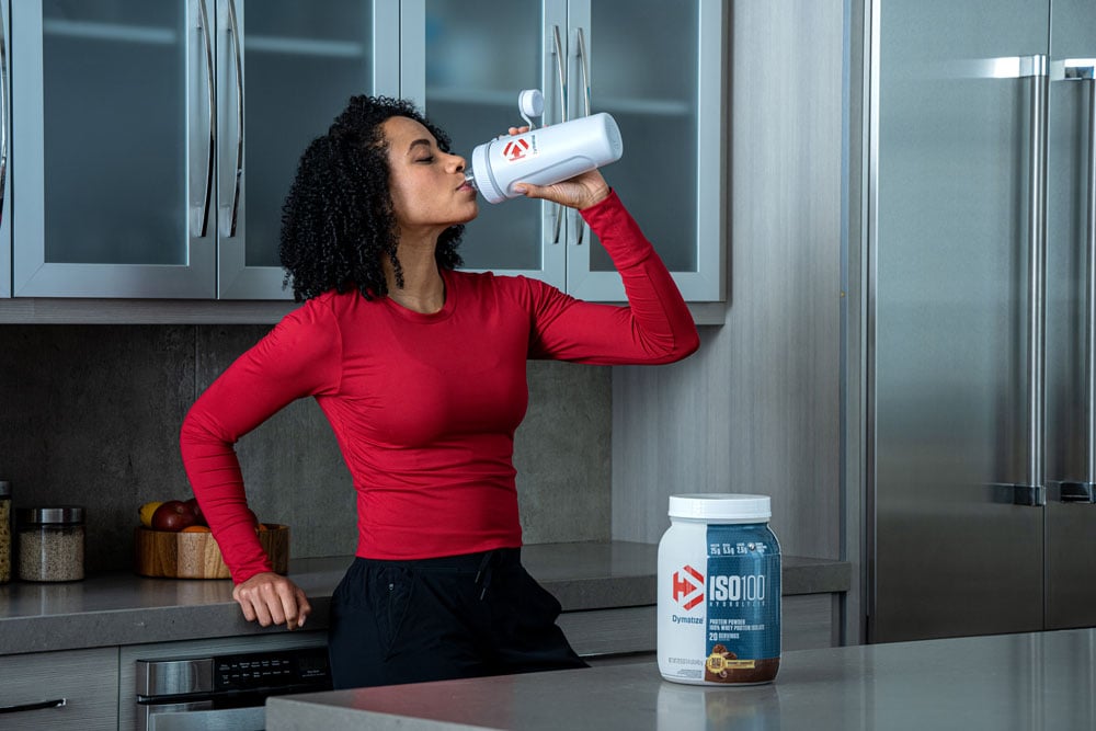 Woman in kitchen drinking out of shaker bottle with container of protein powder on counter.