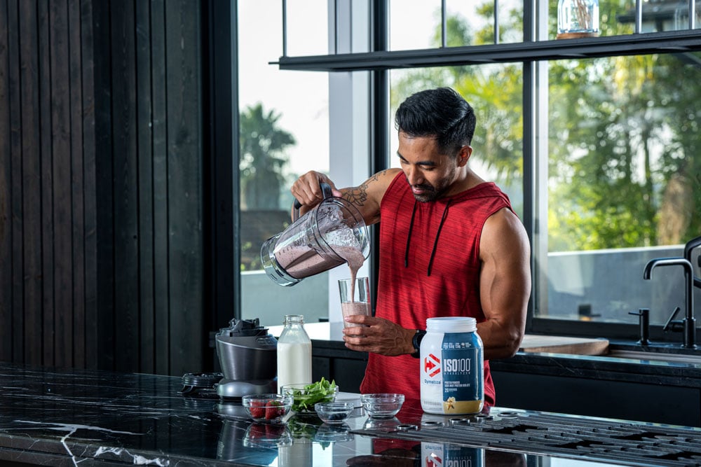 Man in kitchen pouring a protein smoothie into a glass with Dymatize protein powder on counter.