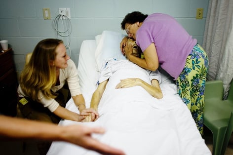 One woman cradles a man's head while he lies in a hospital bed. Another woman holds his hand.