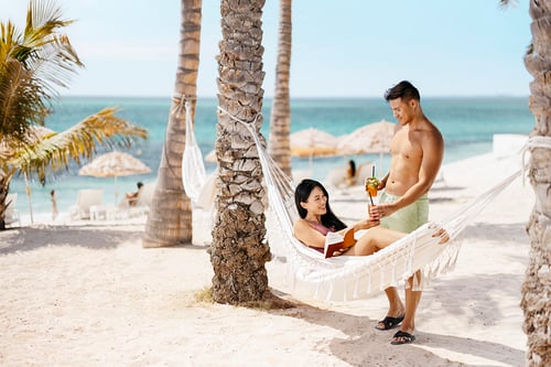 A man hands a woman a drink while she relaxes in a hammock on the beach. Image taken for Abu Dhabi Tourism campaign by photographer Tom Parker.