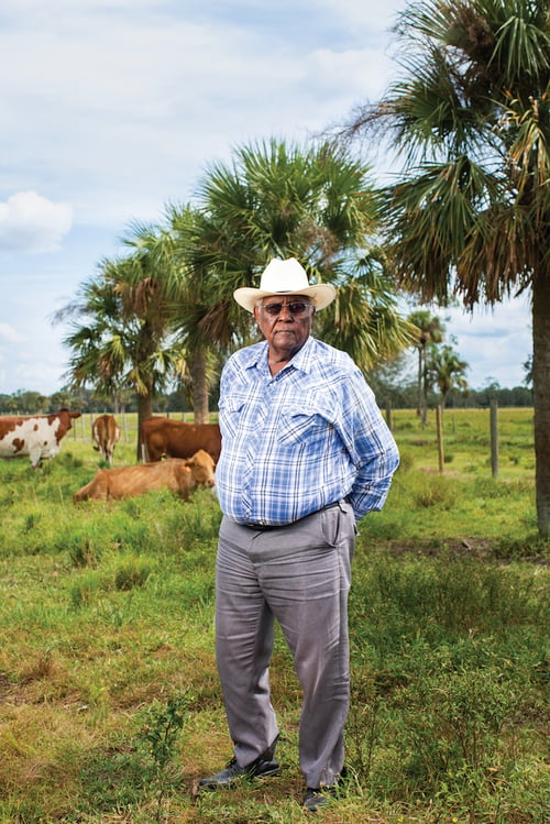 Photo of African American rancher with sunglasses in cowboy hat and blue plaid button-down shirt, standing before cattle and palm trees, by Tampa, Florida-based agricultural photographer Jeremiah Wilson.