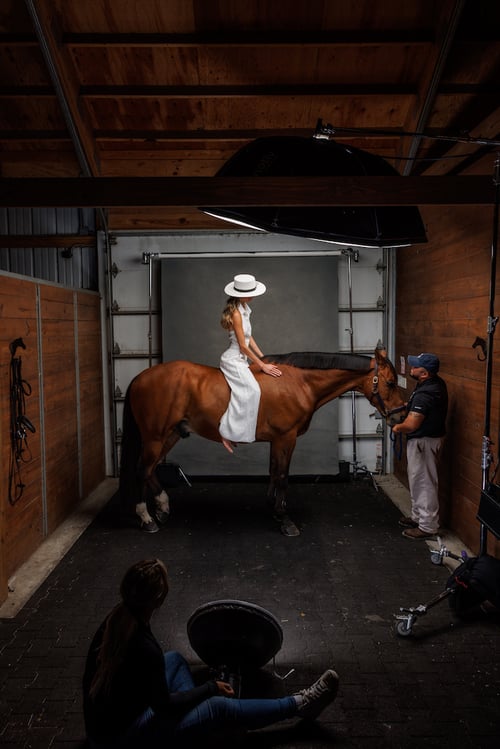 Behind the scenes portrait of talent on horseback in a white onesie, in front of canvas screen, with strobes, horse handler and photographer in foreground.