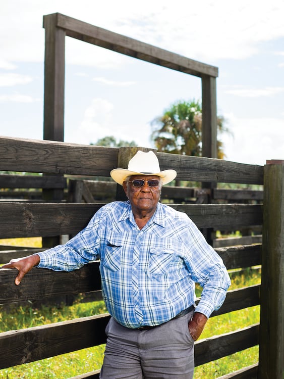 Portrait of African American rancher with sunglasses in cowboy hat and blue plaid button-down shirt, standing before weathered wooden ranch fence, by Tampa, Florida-based agricultural photographer Jeremiah Wilson.