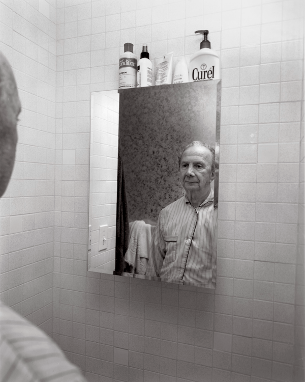 Image of elderly figure in button-down top peering into bathroom mirror with tiled walls, by 2018 grantee Stephen DiRado. 