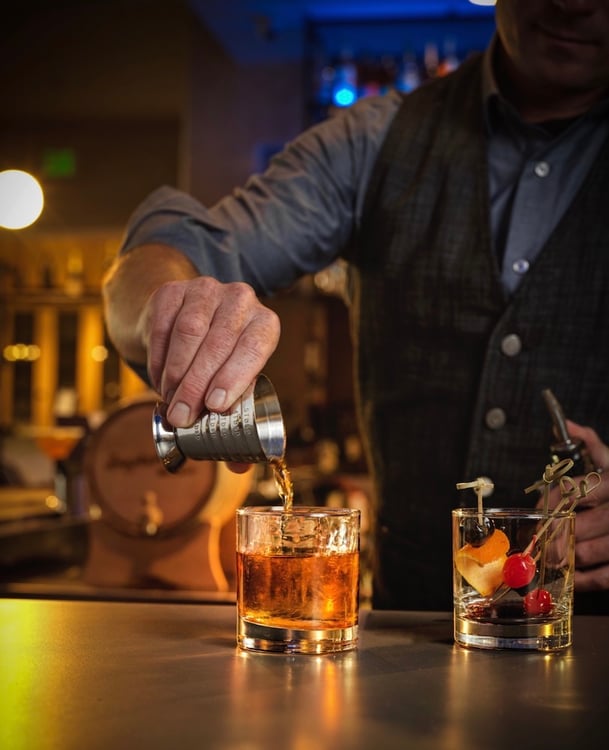 Photo of bartender pouring dark spirits into rocks glass at bar, next to glass of cocktail garnishes, by Seal Beach, California-based food/drink and music/performing arts photographer Eric Hameister.