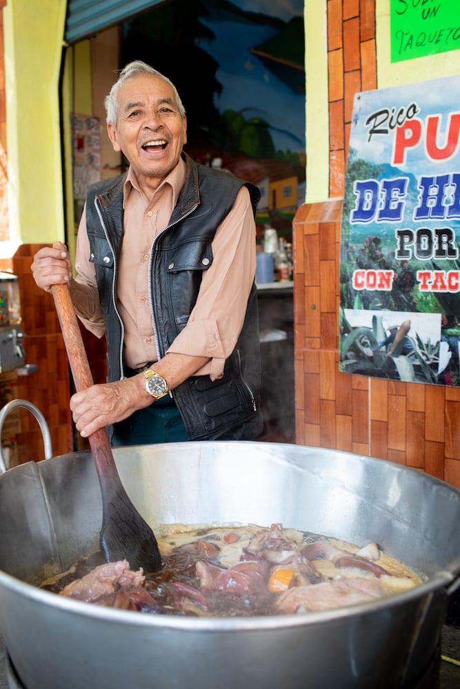 Portrait of man in black leather vest smiling while stirring large stew with wooden paddle, by Mexico City-based travel photographer Andrew Reiner. 