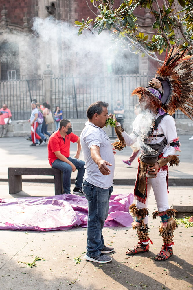 Image of medicine man in traditional costume holding green herbs and a smoking chalice to a close-eyed man's nose, standing arms spread on street, by Mexico City-based travel photographer Andrew Reiner. 