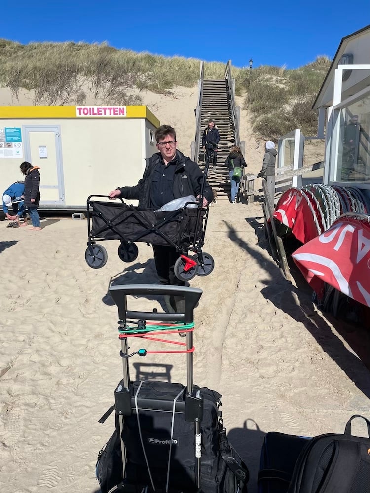 Behind the scenes image of photo assistant carrying photo equipment onto Dutch beach on clear day, by Tilburg, Netherlands-based portrait photographer René van der Hulst.