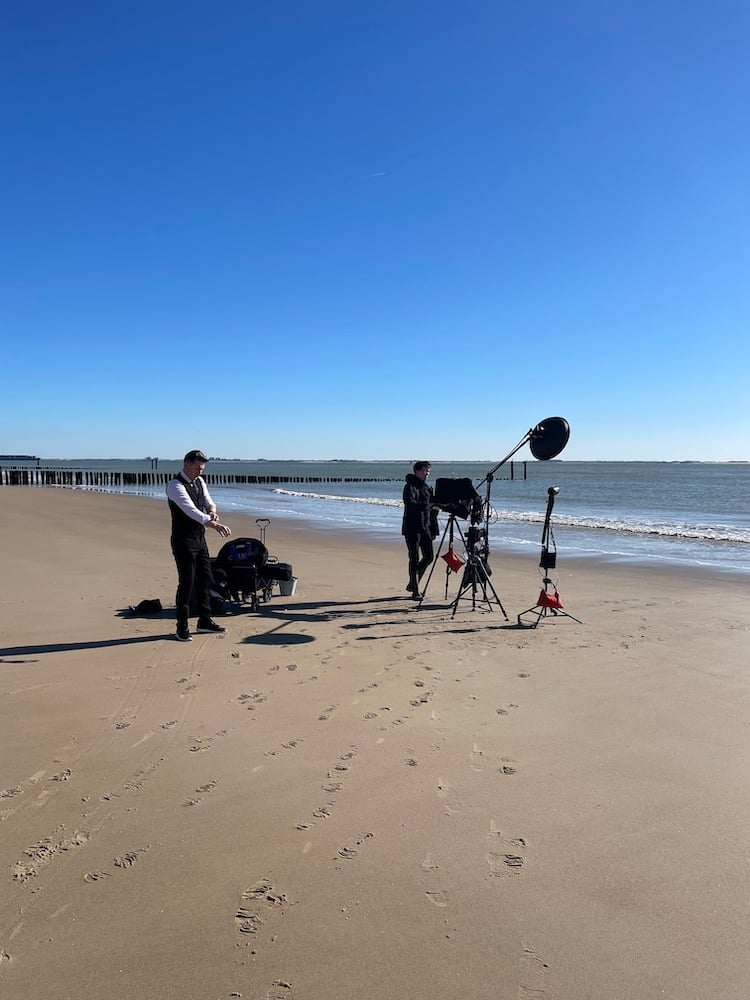Behind the scenes photo of subject in white dress shirt and black vest and pants, and photo assistant with photo equipment on Dutch beach on clear day, by Tilburg, Netherlands-based portrait photographer René van der Hulst.
