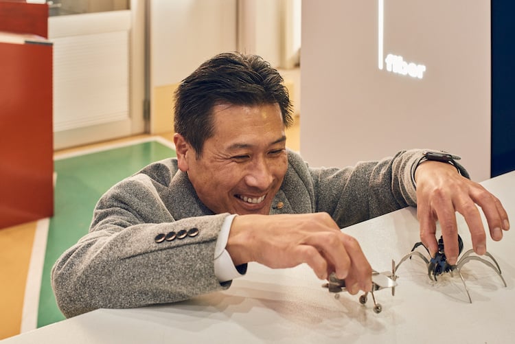 Portrait of adult figure in gray suit jacket, smiling while playing with steel maasa toy airplane and tarantula, by Fukuoka, Japan-based portrait photographer Darien Robertson.