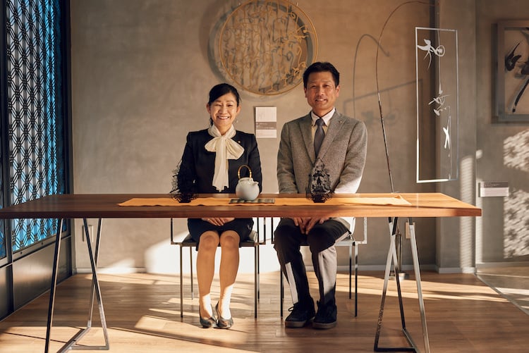 Image of two figures sitting at a wooden table, smiling, with tea pots and maasa decor in background, by Fukuoka, Japan-based portrait photographer Darien Robertson.