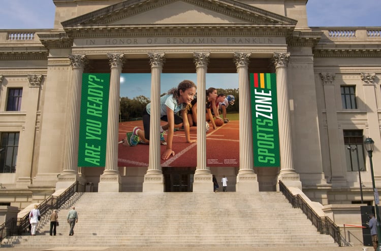 Photo by Colin Lenton of different kinds of athletes of different ages all at the starting line of a racetrack displayed as a banner on the Franklin Institute.