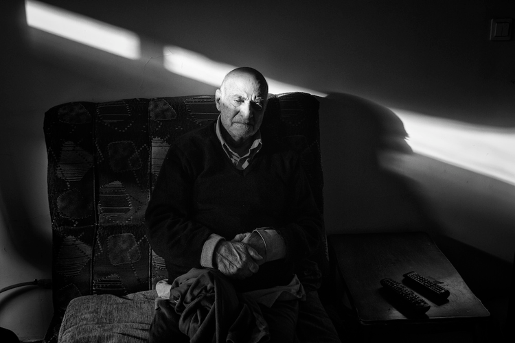 Portrait of bald headed figure seated in arm chair in dark room hands crossed gazing into the near distance, by 2020 grantee Jalal Shamsazaran.