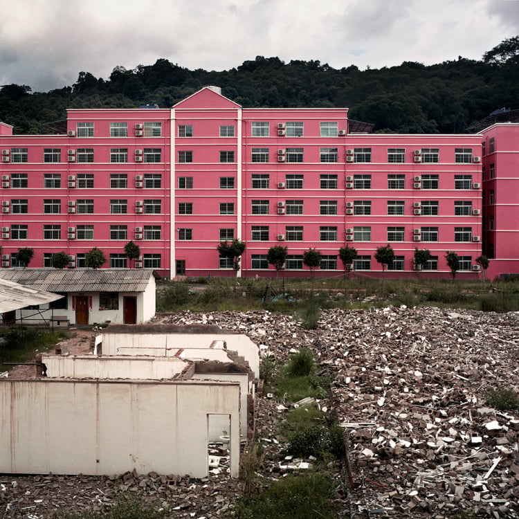 Photo by Ore Huiying of a large, pink apartment building next to a yard of rubble.