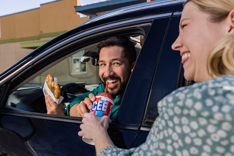 Photo by Zave Smith of a woman handing a man in a car a slurpy and pretzel.