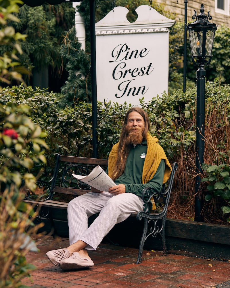 Photo of long-haired bearded figure seated, smiling in green pullover in front of sign for Pine Crest Inn, by Charlotte, North Carolina-based fashion photographer Jackson Ray Petty.