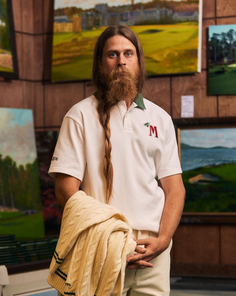 Portrait of long-haired bearded figure in white golf shirt standing before canvas paintings and prints, by Charlotte, North Carolina-based fashion photographer Jackson Ray Petty.