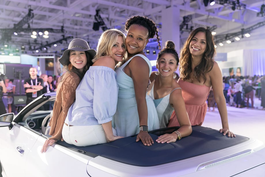 Five young women dressed in pastels sitting on the back of a convertible on a trade show floor captured by Margo Moritz