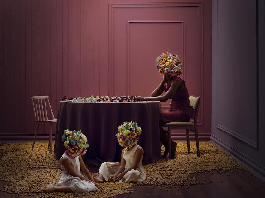 Photo of a woman and two girls by a table, with their heads covered by bouquets of flowers.