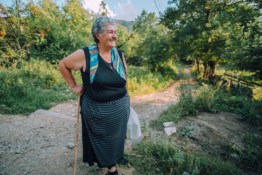 Dimitri Mais' snapshot of an older woman in Georgia, smiling, holding a plastic shopping bag and walking stick