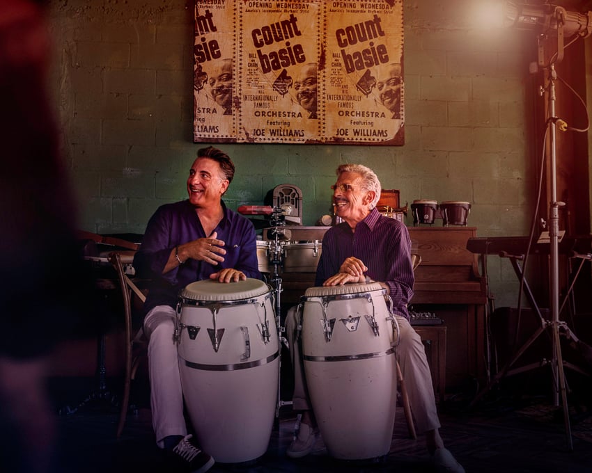 Photo of Andy Garcia with an older man sitting in a room of instruments both playing a Tumbadora drum.