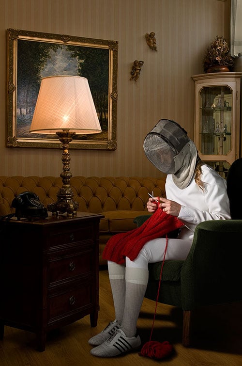 An unusual portrait of a fencing athlete sitting in an armchair, knitting a red garment, in full athletic kit.