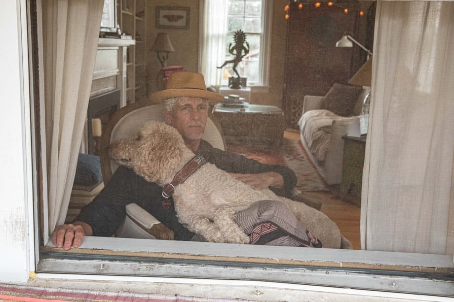 Photo of a man in a tan hat sitting and looking out of his window with his dog.