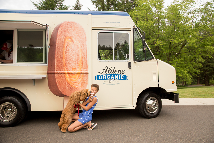 Photo of a woman holding sharing her ice cream cone with her dog in front of an ALden's Ice Cream truck for Alden Ice Cream by John Valls.