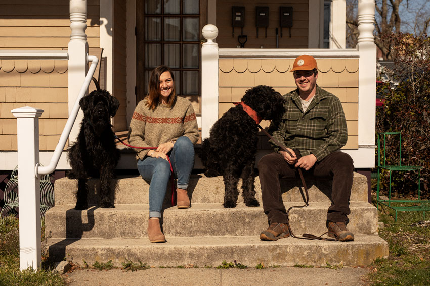 Josh Behan's COVID project Front Porchtrait shows a young man and woman sitting in front of their house with two black dogs