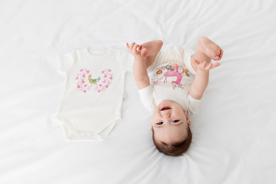Photograph of a baby in Olivia Yves clothing lying on the bed next to a onesie.