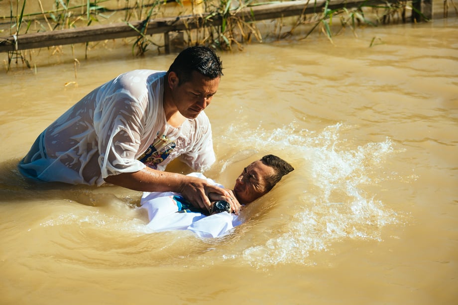 A priest baptizes a pilgrim in the Jordan river in this photo by David Vaaknan