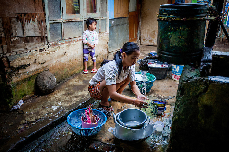 Image of a woman in Indonesia crouching to wash her dishes under water from a public faucet.