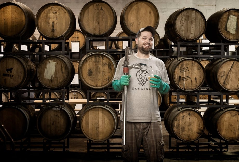Image of an owner at Nightshift viewing posing in front of the barrells their beer ages in, photo by Josh Andrus