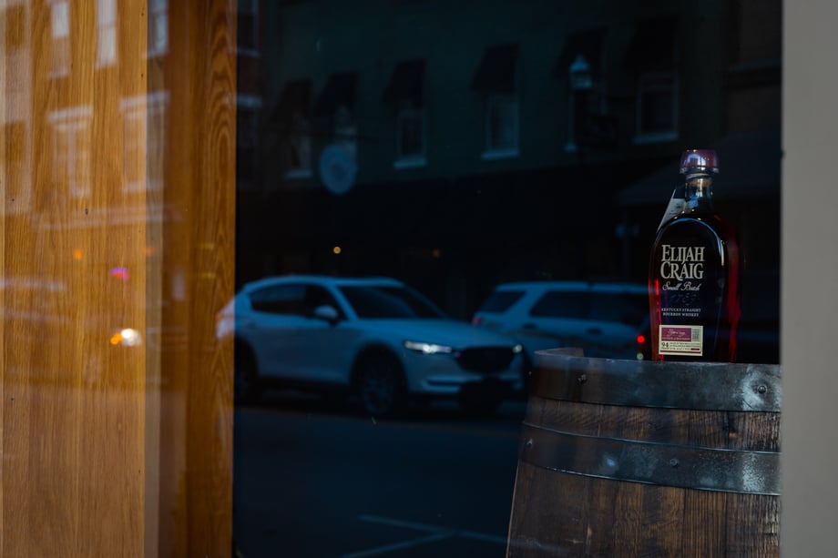 Photo by Matthew Allen of a bottle of Elijah Craig bourbon through a storefront window where you can see a Mazda in the reflection