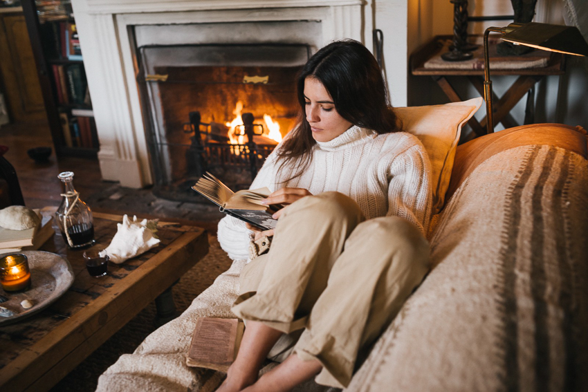 Portrait of woman sitting by fireplace reading a book by Photographer Aaron Greene for We Took to the Coast