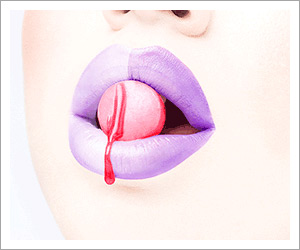Aj Raina's closeup beauty image of a mouth with purple lipstick and a piece of candy 