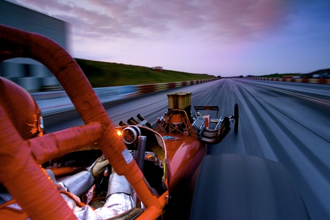 A red dragster is shot by Dom Romney from near the driver's seat while on the raceway