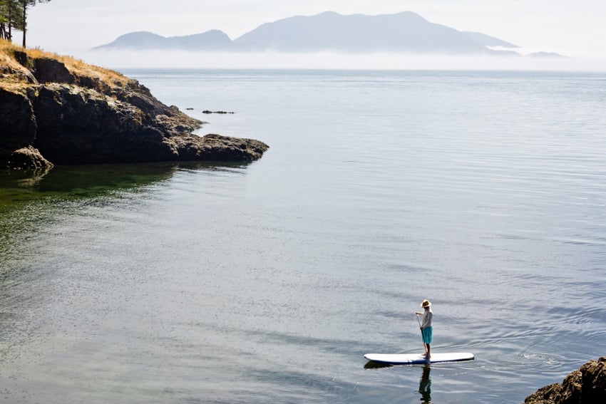 A lone paddle boarder making small waves next to a rocky shoreline.