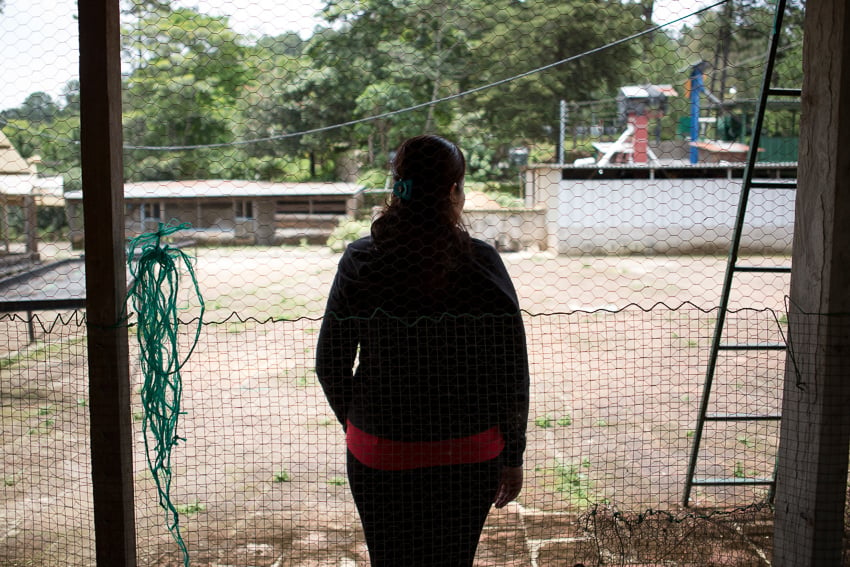 Photo of a woman behind a fence looking out into a yard published on The Intercept taken as part of an IWMF Adelante reporting fellowship by Alicia Vera