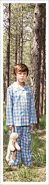 A young boy stands in a forest in blue-checkered pjs while holding a teddy bear by James Ellerker