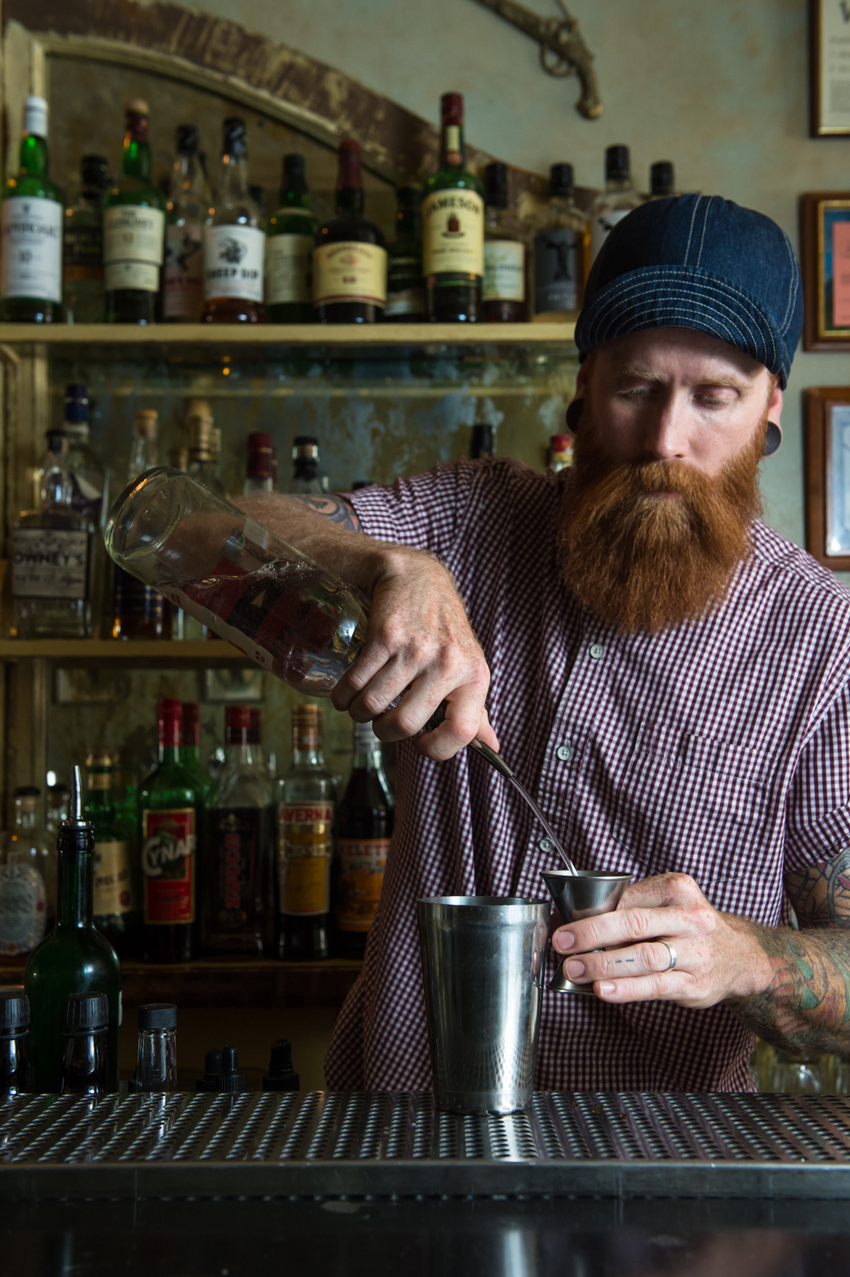 A tattooed bartender pours liquor into a shot glass as he prepares a cocktail, photo by Jennifer May. 