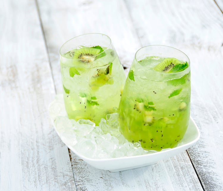 Two glasses of an icy cocktail containing kiwi and mint leaves, sitting in a bowl of ice chips