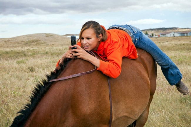 A young woman lying on top of a horse, staring at her cellphone 