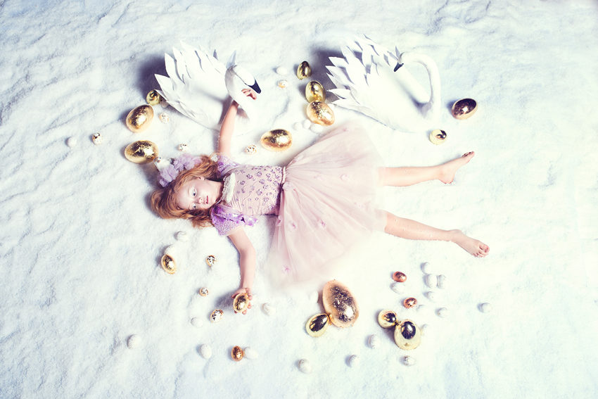 A girl lying in the snow with paper swans and golden eggs, photograph by Megan Dendinger