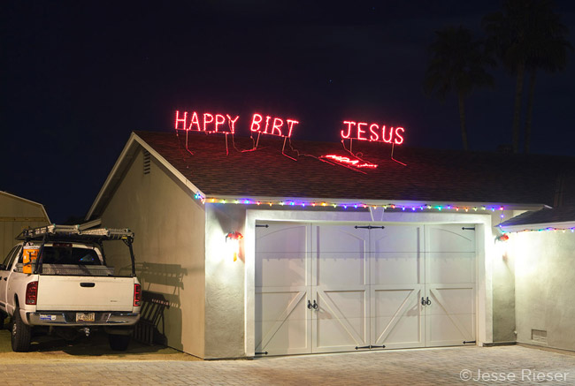 A red neon sign that reads "Happy Birt Jesus" on a house roof shot  by Phoenix-based conceptual photographer Jesse Rieser